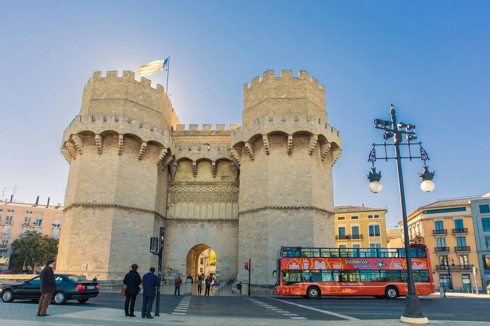 Ιστορική πέτρινη πύλη με δύο στρογγυλούς πύργους κάτω από ένα γαλάζιο ουρανό. Τουρίστες και ένα κόκκινο λεωφορείο για τα αξιοθέατα σε πρώτο πλάνο.