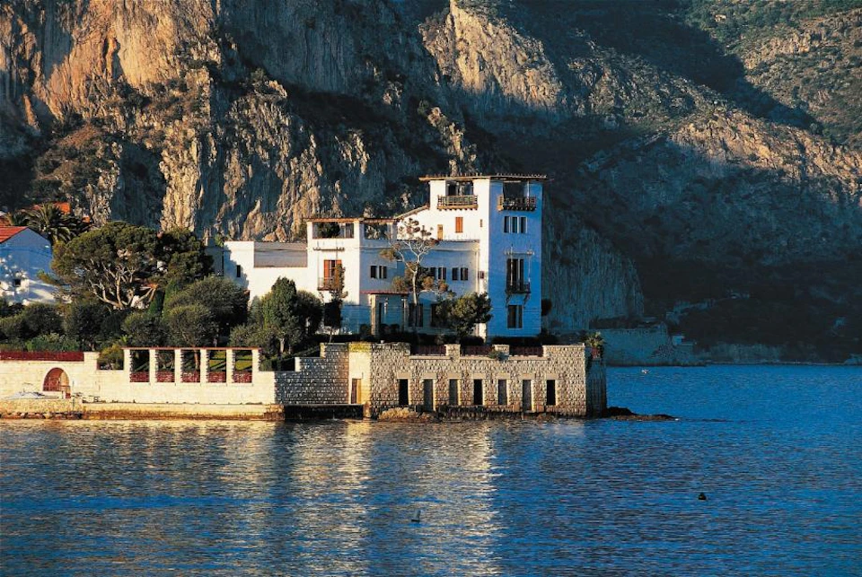 White building with multiple balconies and terraces overlooking a body of water, set against a rocky hillside.