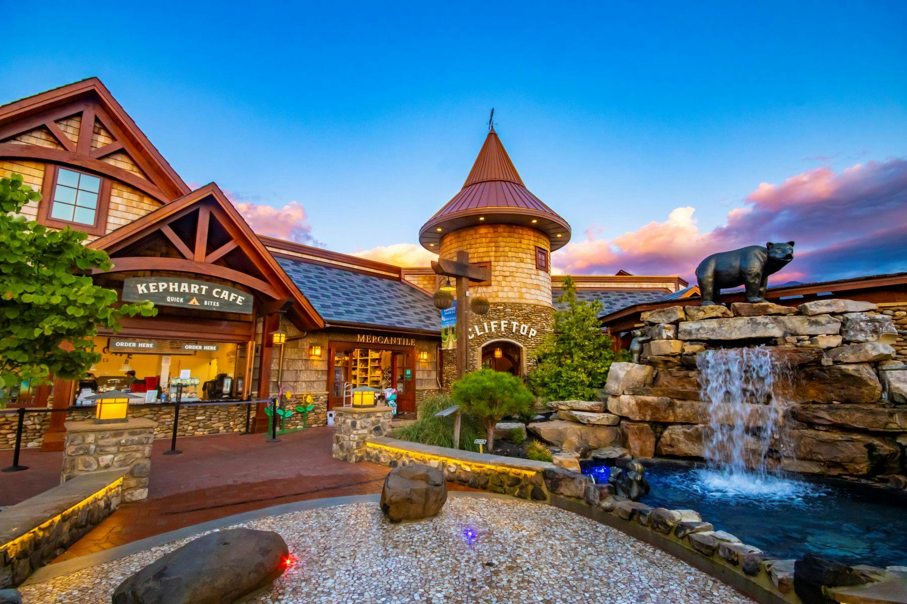 A rustic-themed cafe and a stone tower near a waterfall display under a vibrant sunset sky.