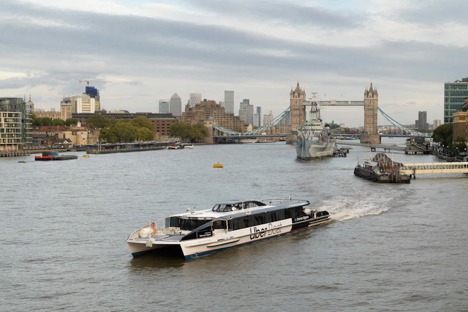 A modern ferry labeled "Uber Boat" travels on a river with Tower Bridge and city buildings in the background.