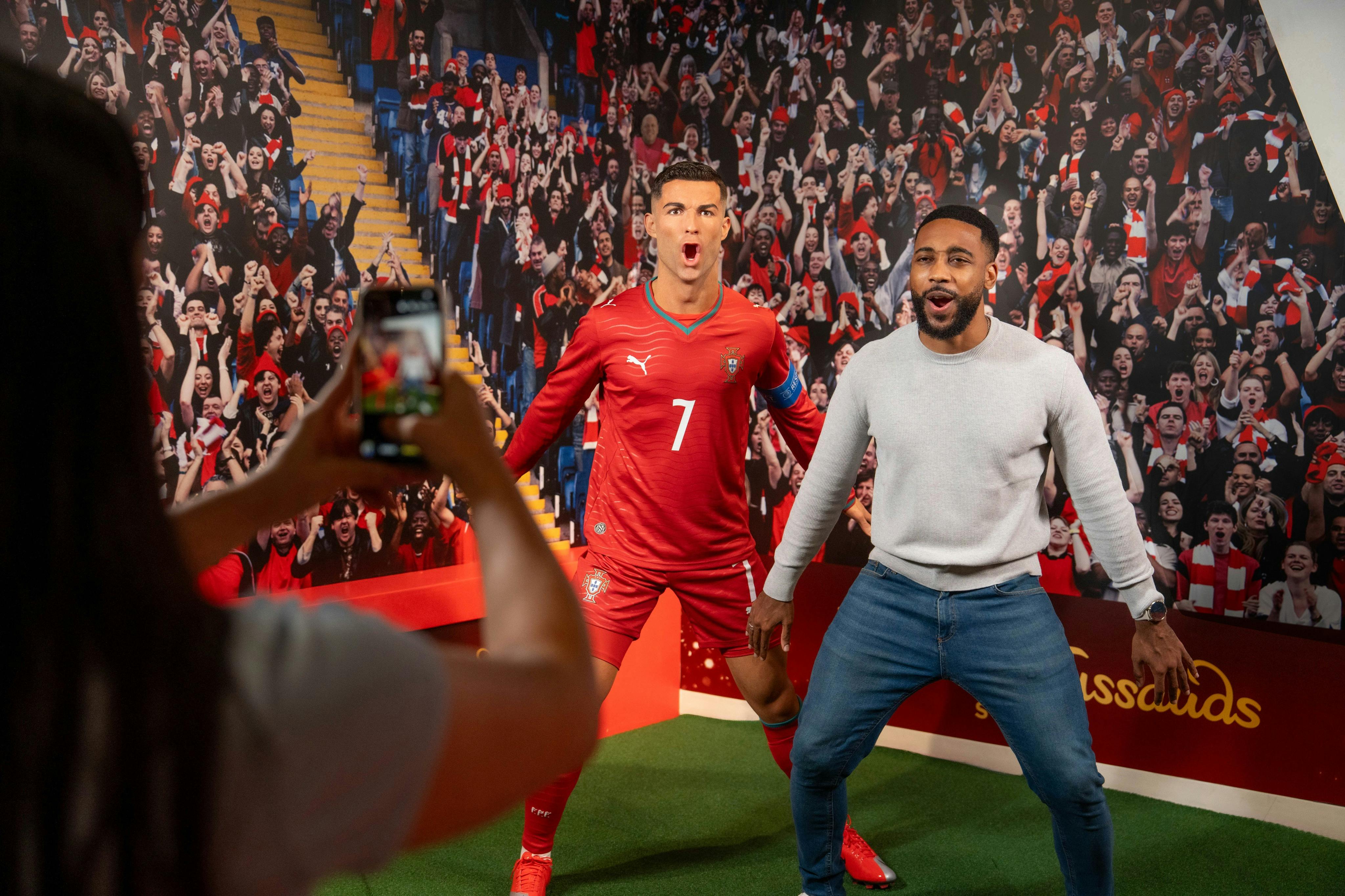 A man poses next to a wax figure of a soccer player in a red uniform, with a crowd backdrop in a museum setting.