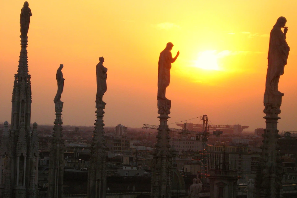 Three statues stand atop stone columns against a cityscape backdrop at sunset, with a clear orange sky.