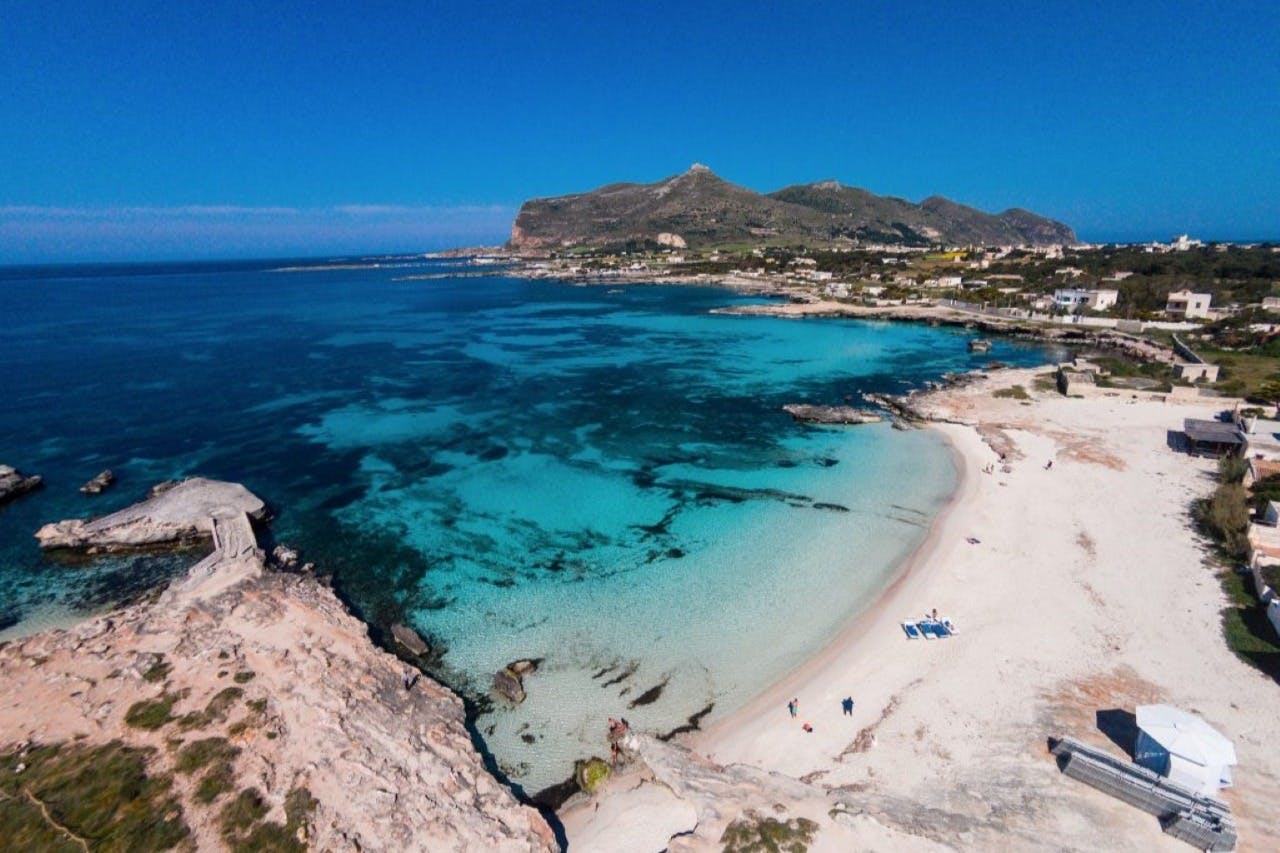 Aerial view of a sandy beach with turquoise waters, rocky shoreline, a few people, and distant mountains under a clear blue sky.