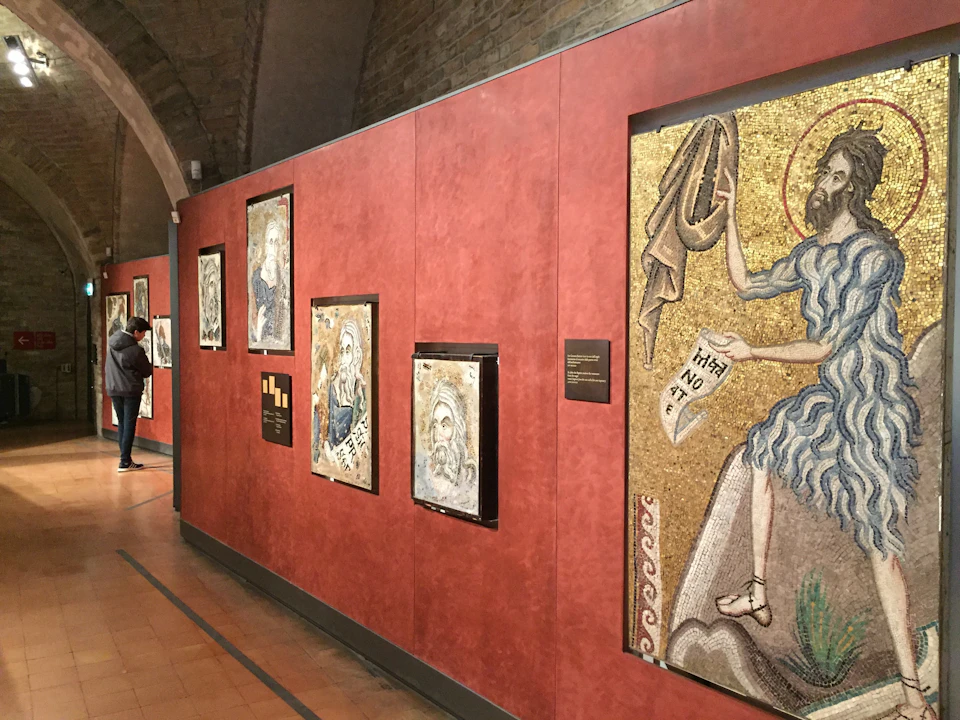 Museum hallway with red walls displaying framed mosaics of religious figures. A person admires the art from a distance.