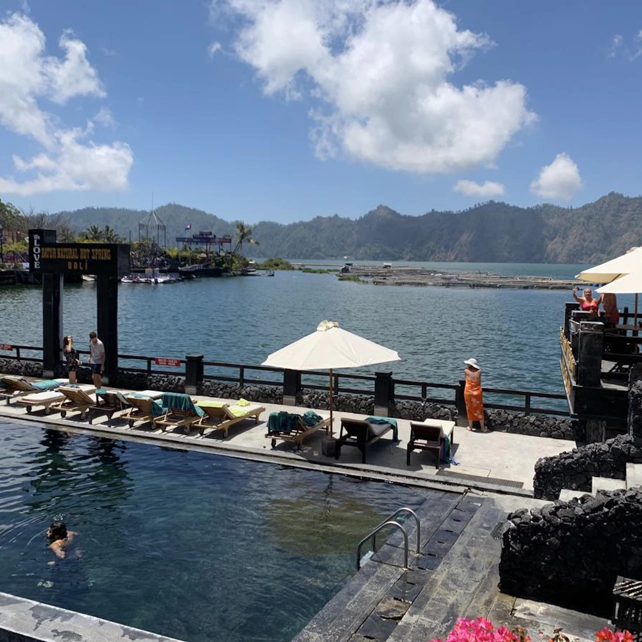 A scenic poolside area with lounge chairs, umbrellas, and people by a lake, with mountains and clouds in the background.