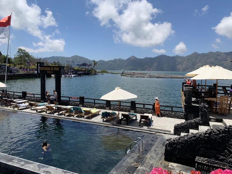 A poolside area with lounge chairs and umbrellas near a lake, surrounded by mountains under a partly cloudy sky.