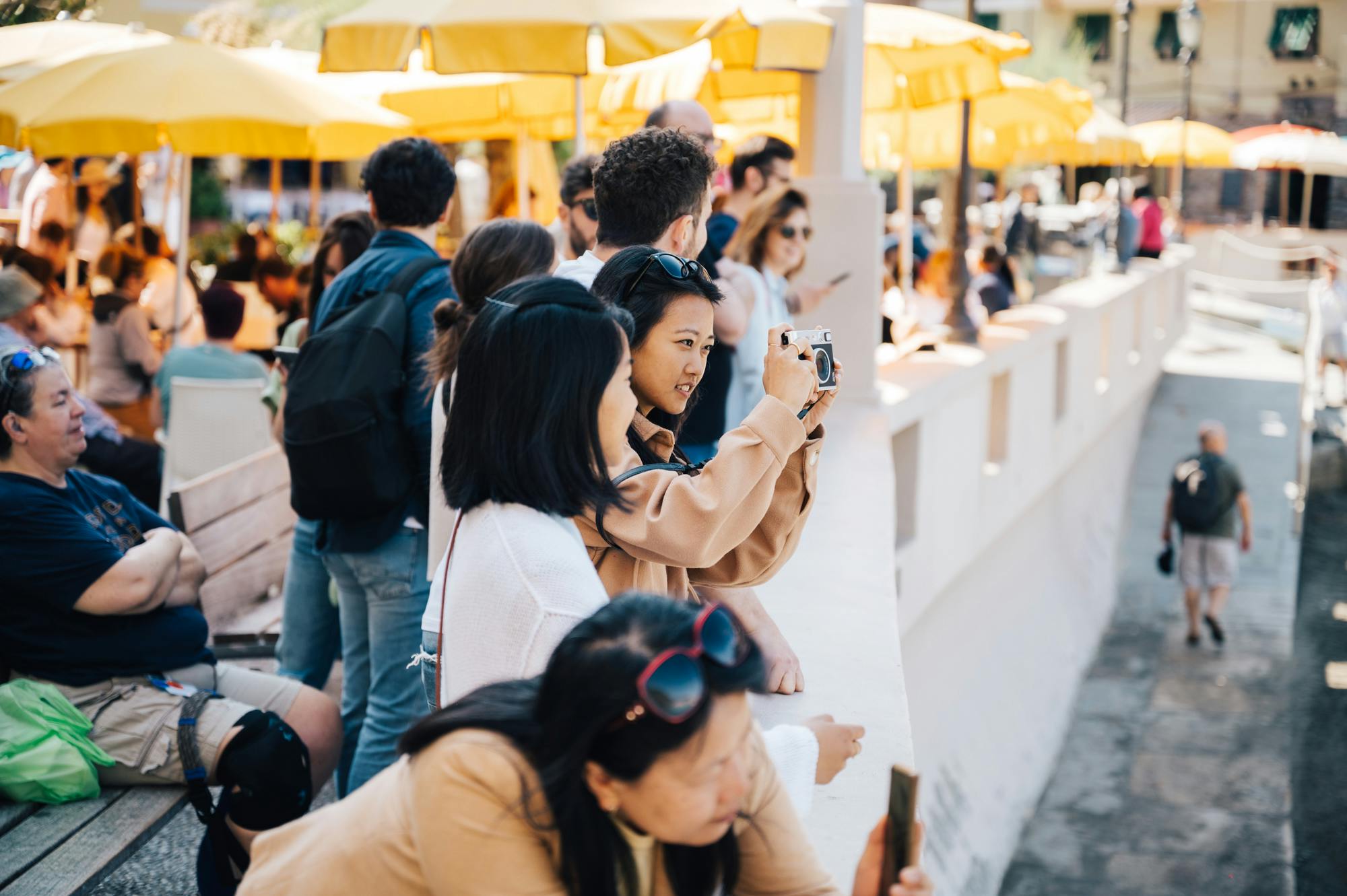 人们在户外聚集在黄色的雨伞下。 一名女性对焦相机，而其他人则坐在或站在附近。