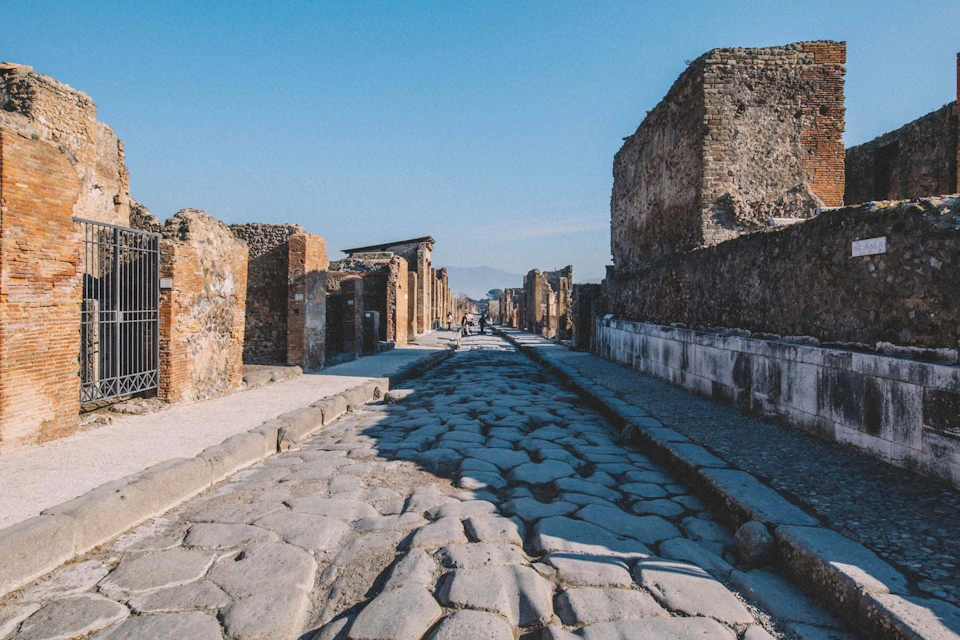 A cobblestone street in an ancient city with weathered stone buildings on both sides under a clear blue sky.