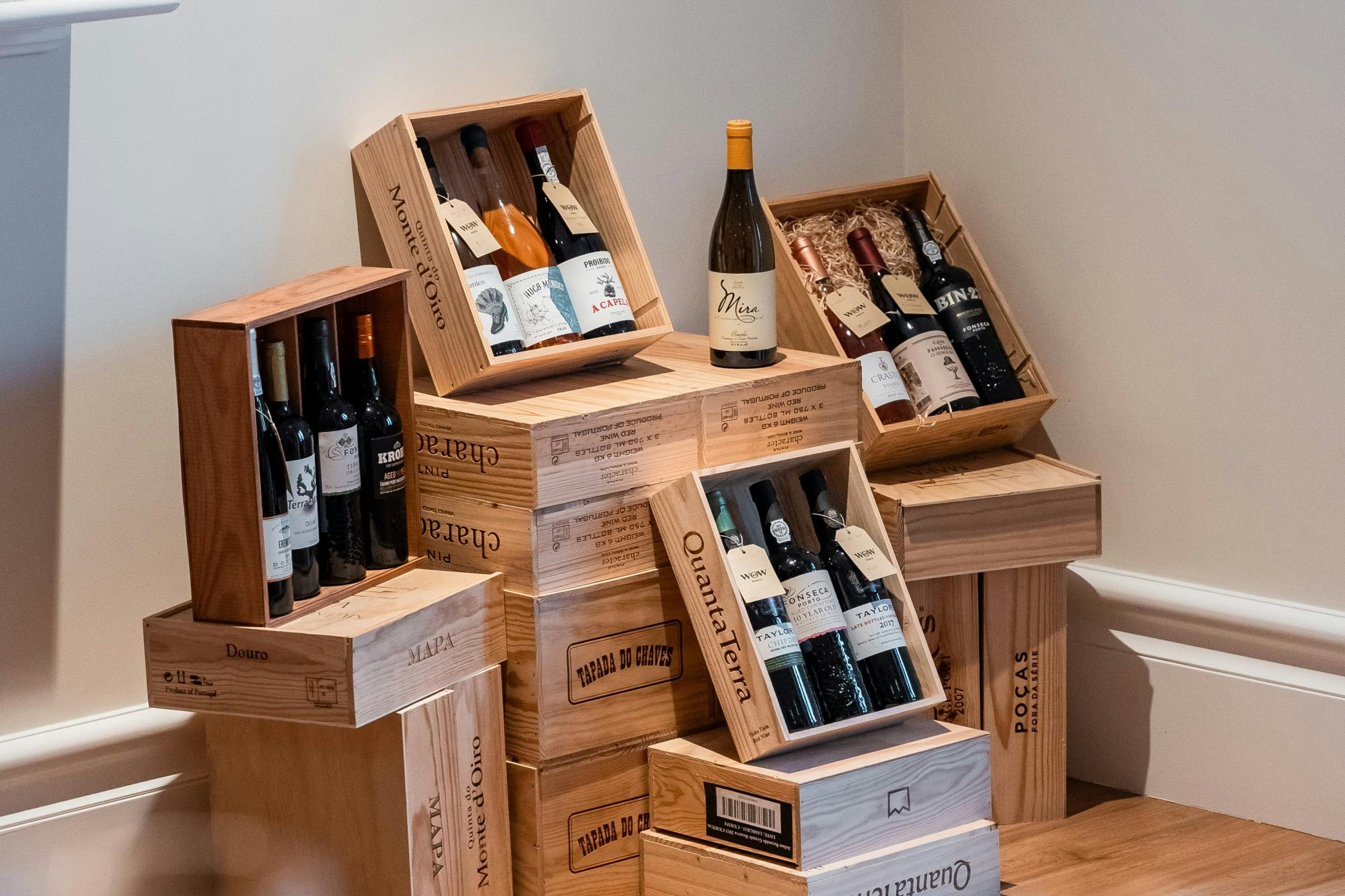 Wooden wine crates, some open to display bottles of wine, are stacked against a beige wall on a wooden floor.
