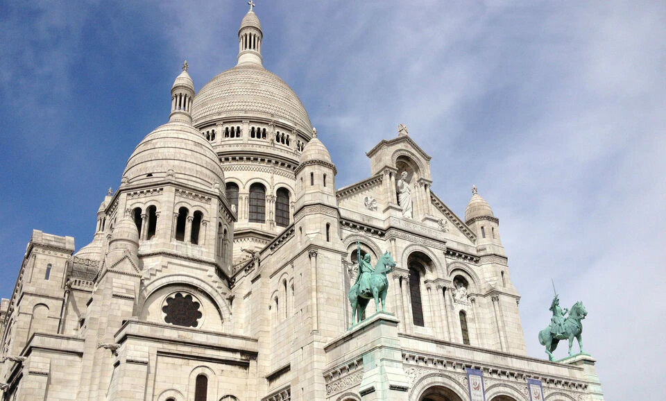 White stone basilica with multiple domes and a statue of a rider on horseback in front, set against a partly cloudy sky.