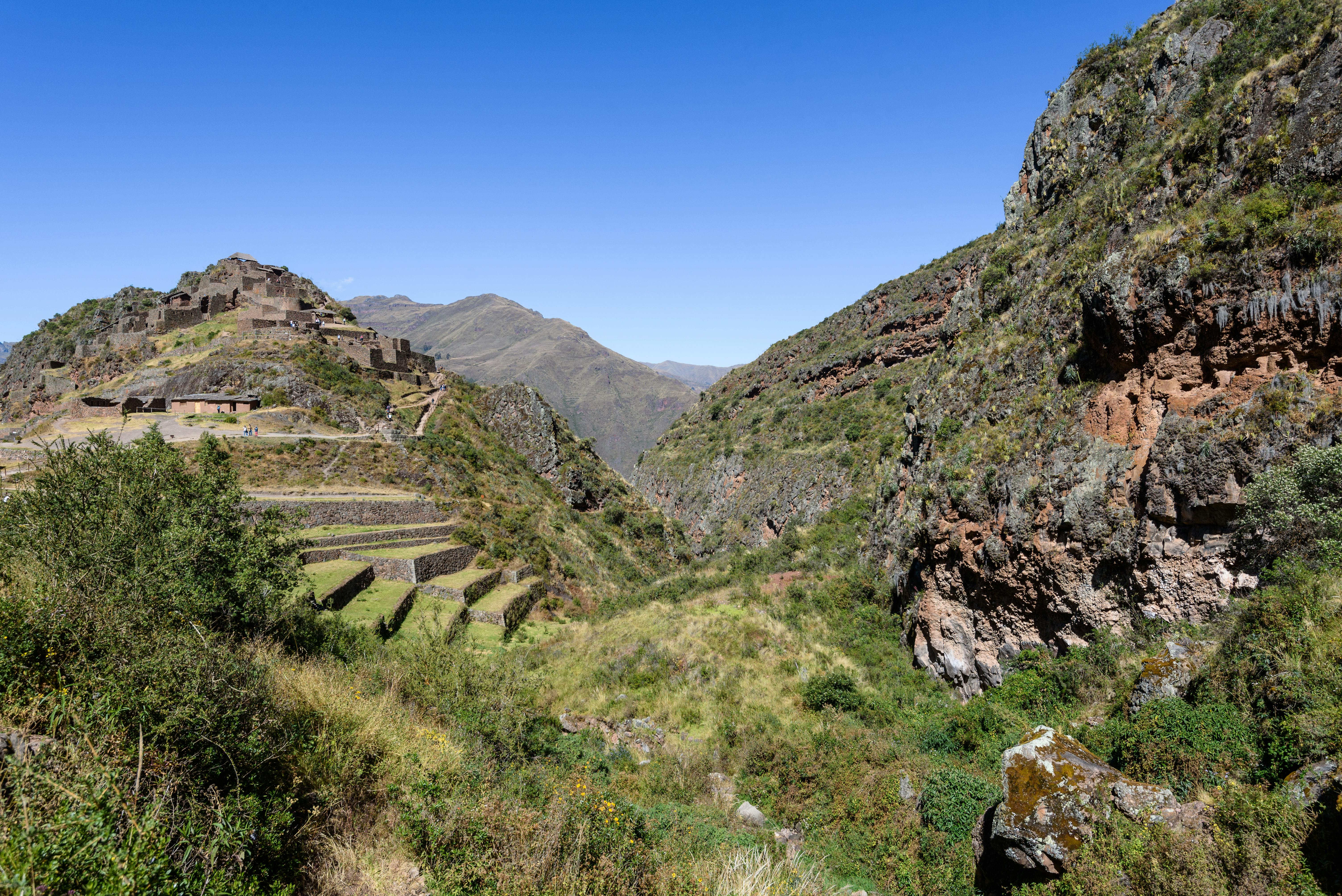 Stone ruins and terraced fields atop a hill, surrounded by steep, green valleys under a clear blue sky.