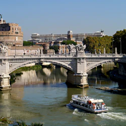 En båt kryssar på en flod under en utsmyckad stenbro med en stor historisk byggnad och träd i bakgrunden.