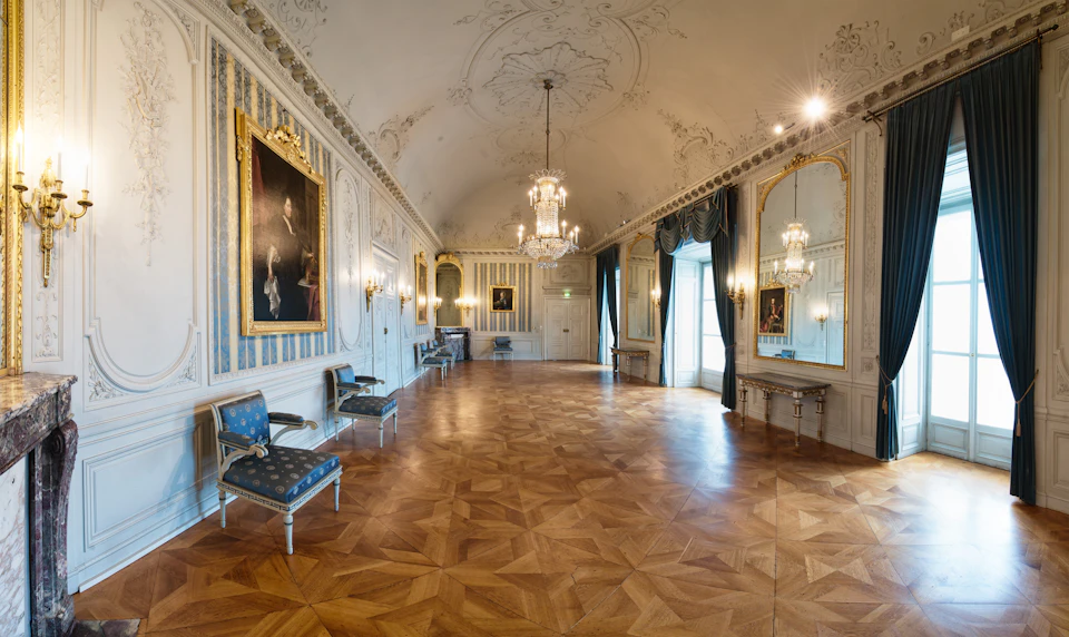 Elegant room with wooden flooring, chandeliers, large mirrors, paintings, and ornate molding. Blue armchairs line the walls.