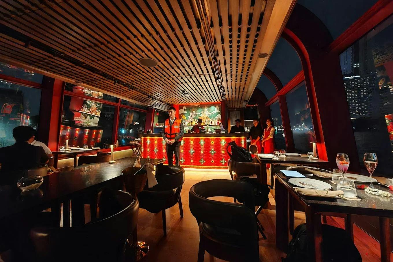 A warmly lit restaurant interior with a wooden ceiling and red accents, featuring a bar and people standing behind it.