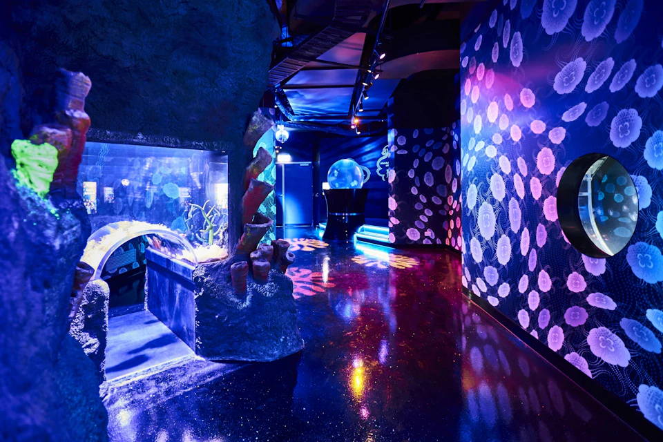 A vibrant aquarium exhibit with illuminated tanks, coral decorations, and jellyfish patterns on the walls under blue lighting.