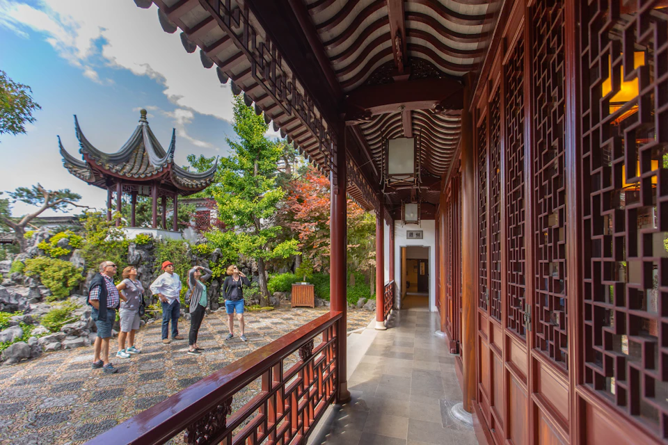 People taking photographs in a traditional Chinese garden with a tiled pavilion, vibrant trees, and a red wooden corridor.