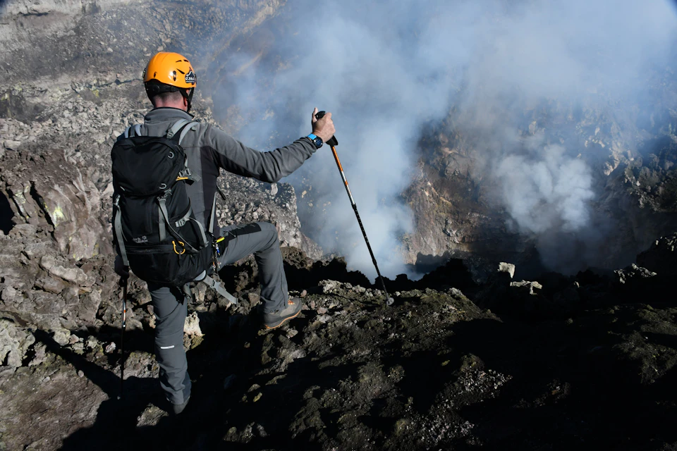 Person in climbing gear and helmet stands on rocky terrain, holding a trekking pole, overlooking a smoky, mountainous area.