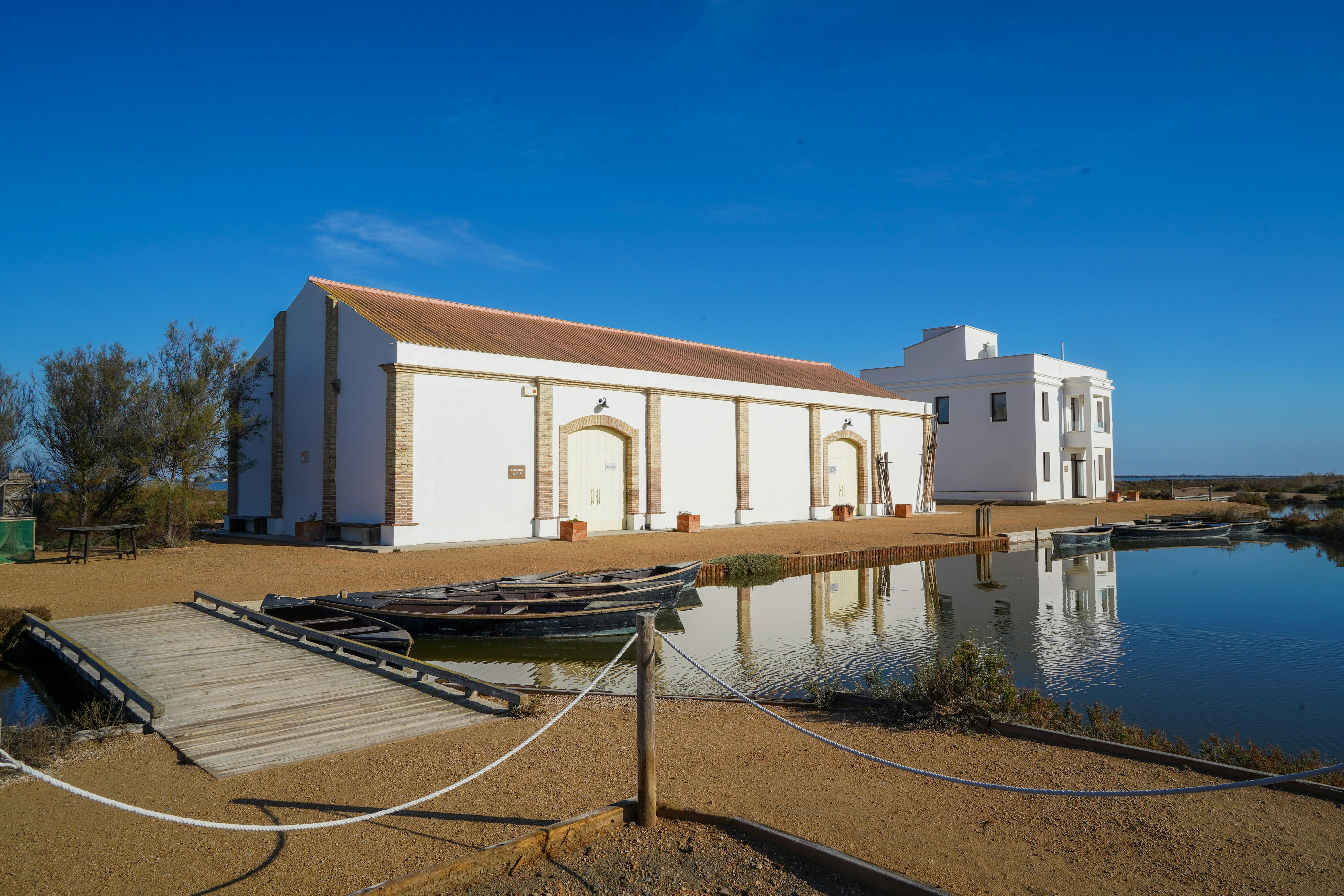 Bílá budova s klenutými dveřmi a okny vedle reflexního rybníka se dvěma malými čluny zakotvenými u dřevěné cesty.