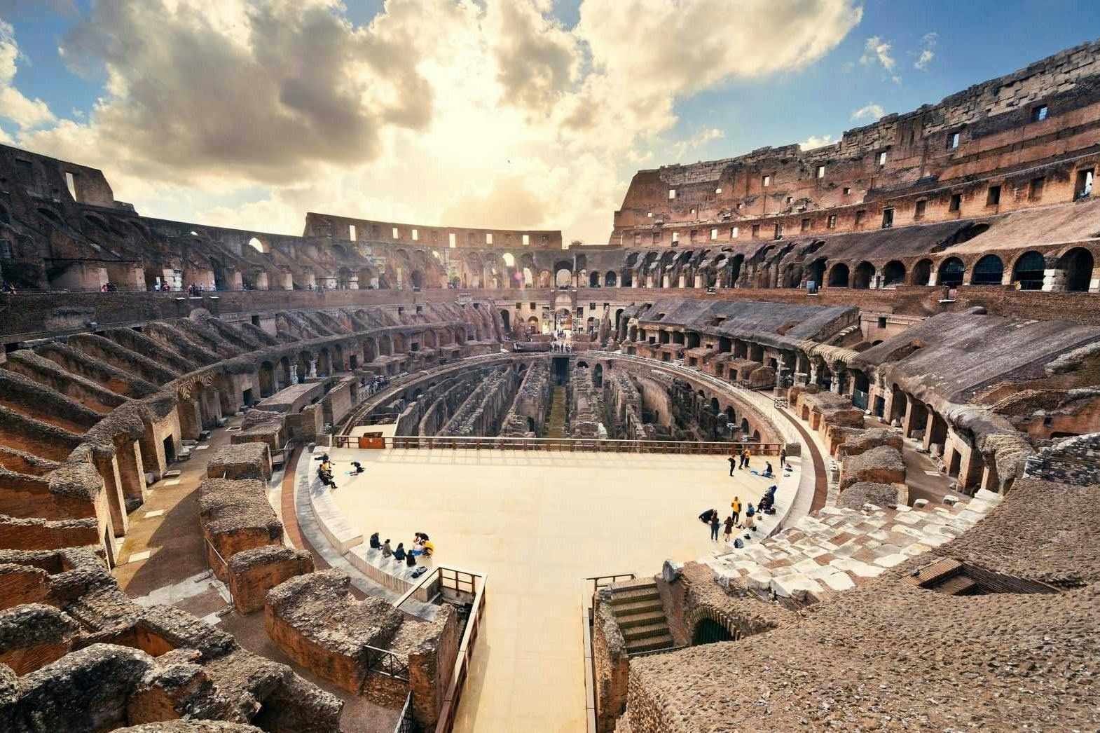 Vista interna del Colosseo a Roma sotto un cielo parzialmente nuvoloso, con i visitatori che esplorano le rovine storiche.