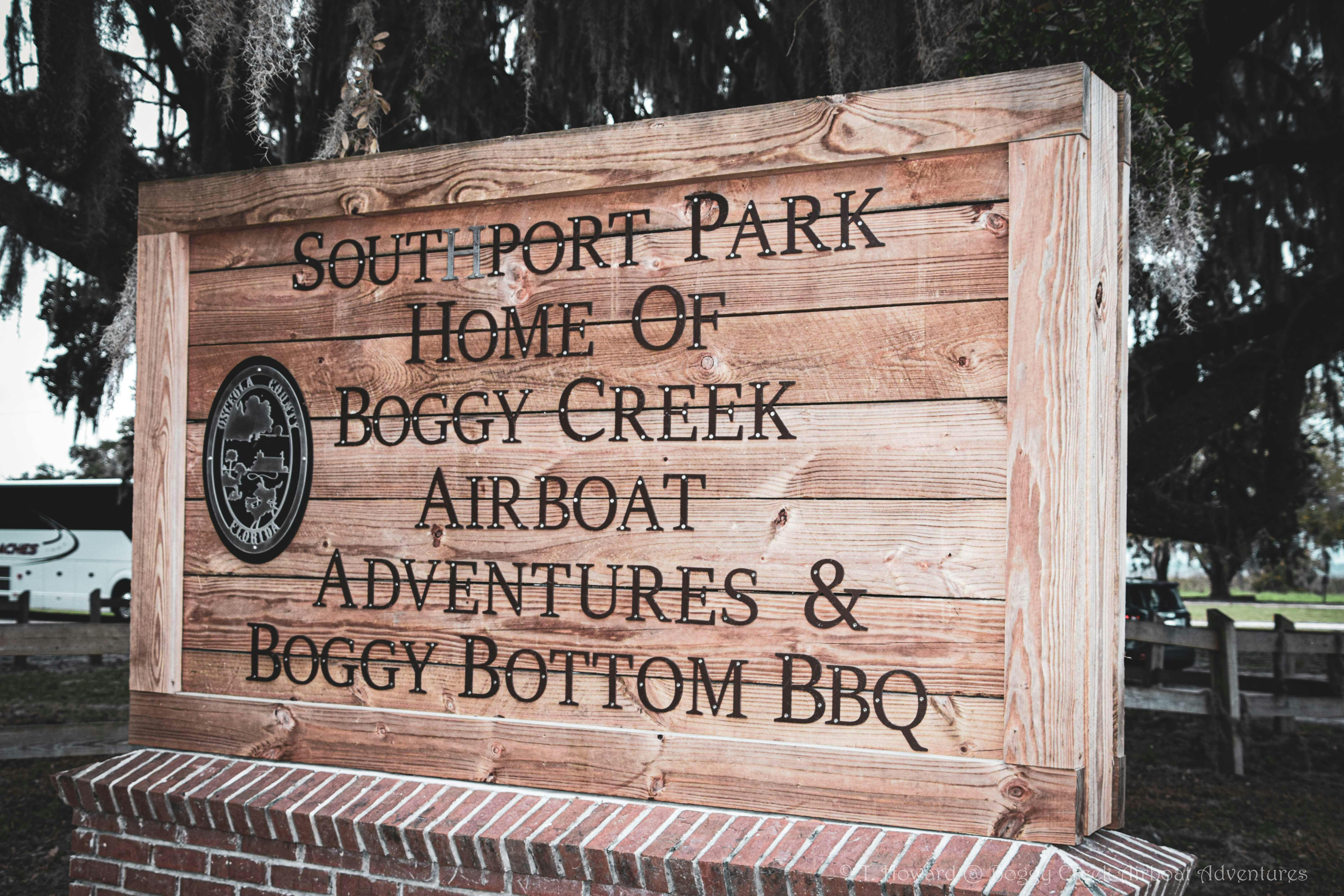 Ξύλινη επιγραφή που γράφει «Southport Park Home of Boggy Creek Airboat Adventures & Boggy Bottom BBQ» με εγχάρακτη σφραγίδα στα αριστερά.