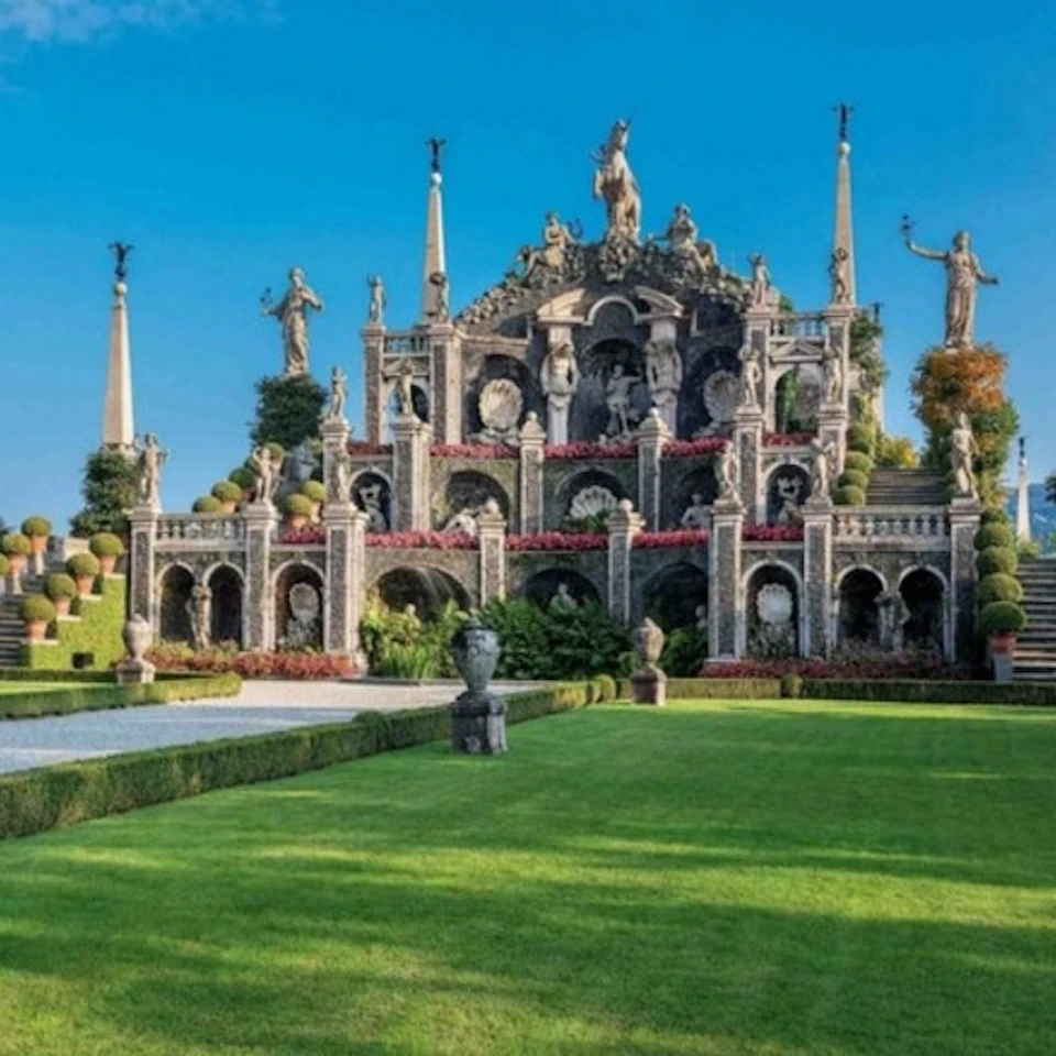 Ornate garden with manicured lawns, statues, tiered fountains, and decorative arches against a clear blue sky.