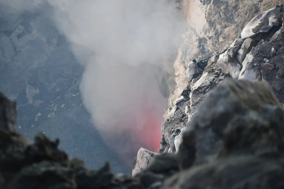 A rocky, geothermal landscape with smoke rising from a fissure, suggesting volcanic activity. A hint of red glow is visible through the smoke.