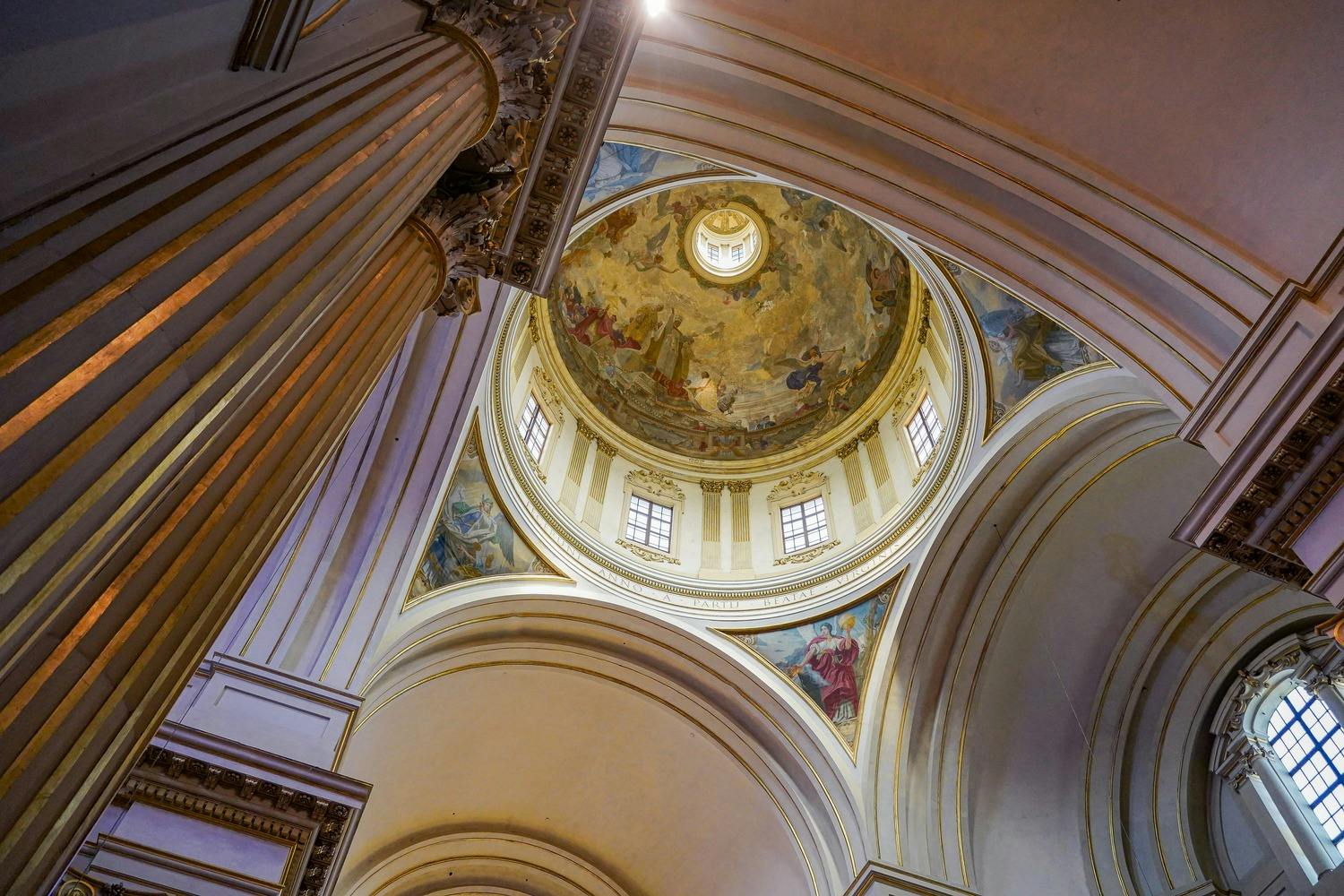 Intricately painted domed ceiling of a cathedral with ornate columns and arched windows, viewed from below.