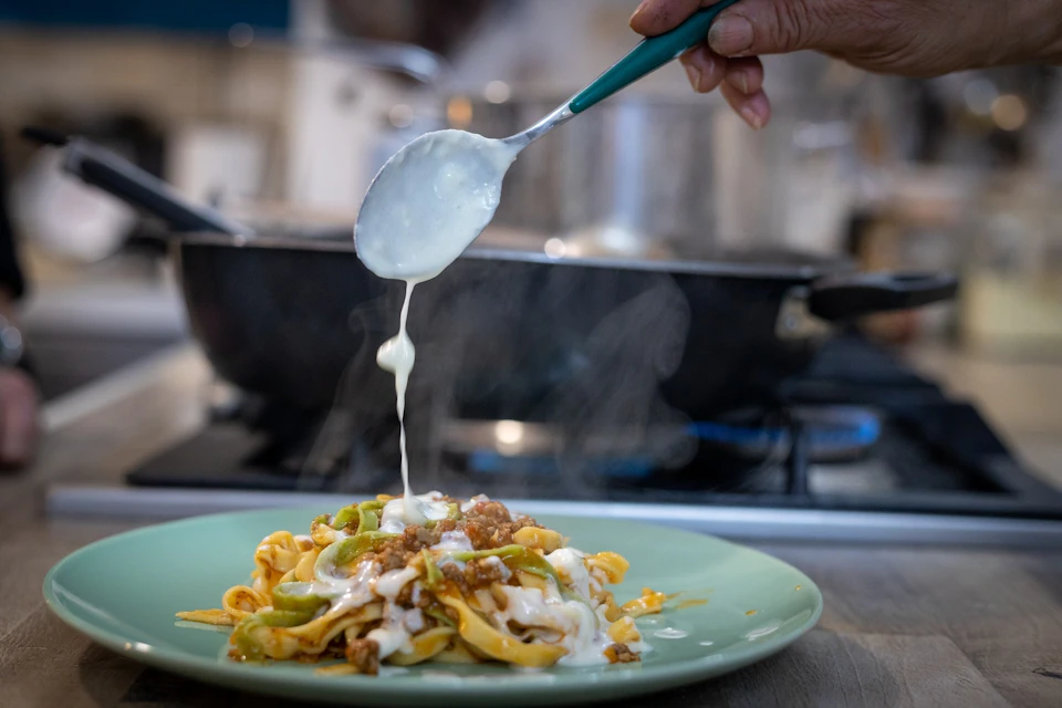 A hand pours creamy sauce over steaming pasta with vegetables and meat on a green plate in a kitchen setting.