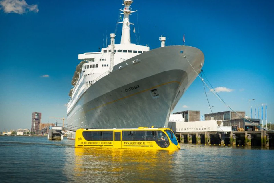 A large ship named "Rotterdam" docked near a yellow amphibious tour bus on water, under a clear blue sky.