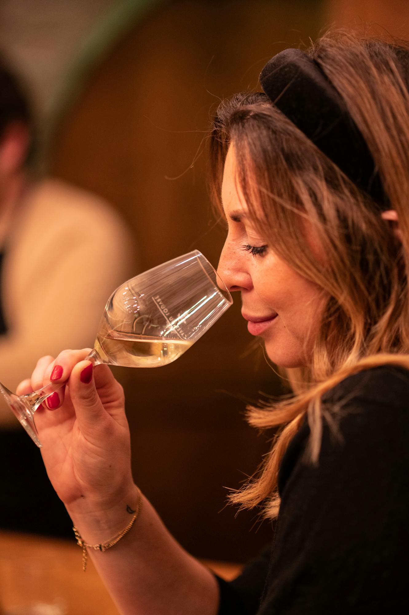 Женщина с длинными волосами, держащая бокал белого вина, пахнущий его ароматом, в тепло освещенном помещении.