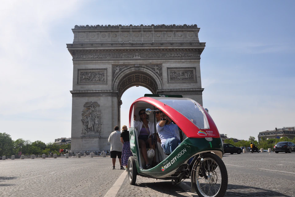 A pedicab with passengers in front of the Arc de Triomphe in Paris. Other pedestrians are visible in the background.