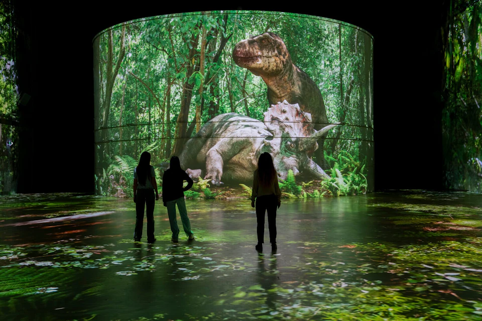 Three people stand in front of a large digital display showing a forest scene with two dinosaurs, a T. rex and a triceratops.