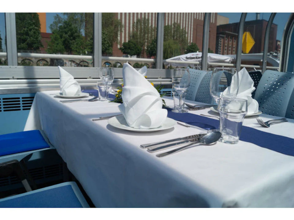 An outdoor dining table set with white cloth, folded napkins, wine glasses, and cutlery. Urban buildings visible in the background.