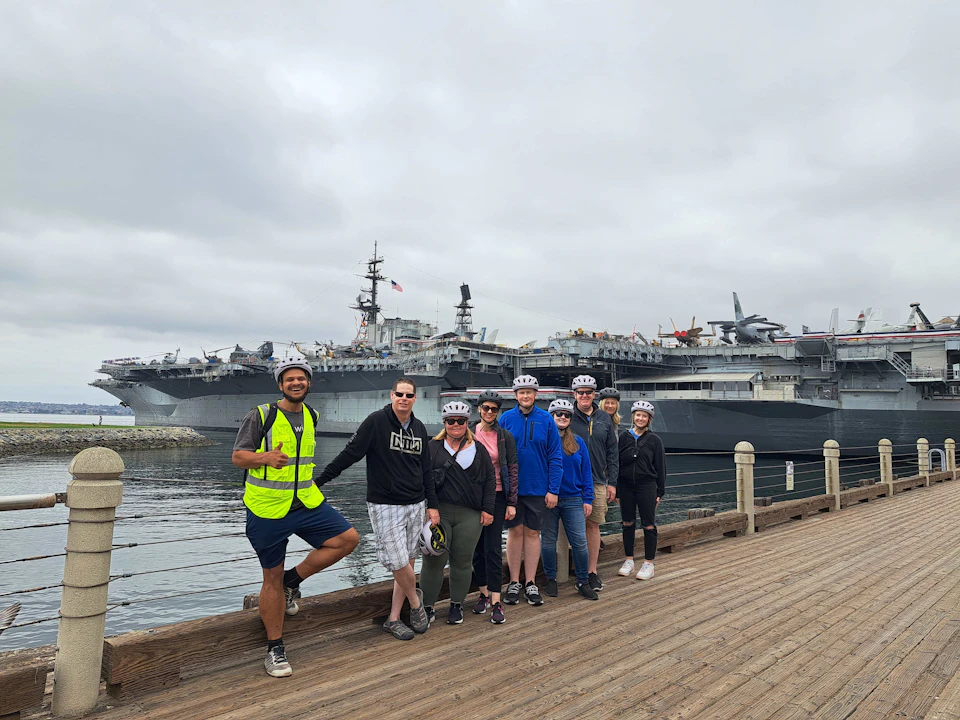 A group of people wearing helmets pose on a boardwalk with a large aircraft carrier docked in the background.