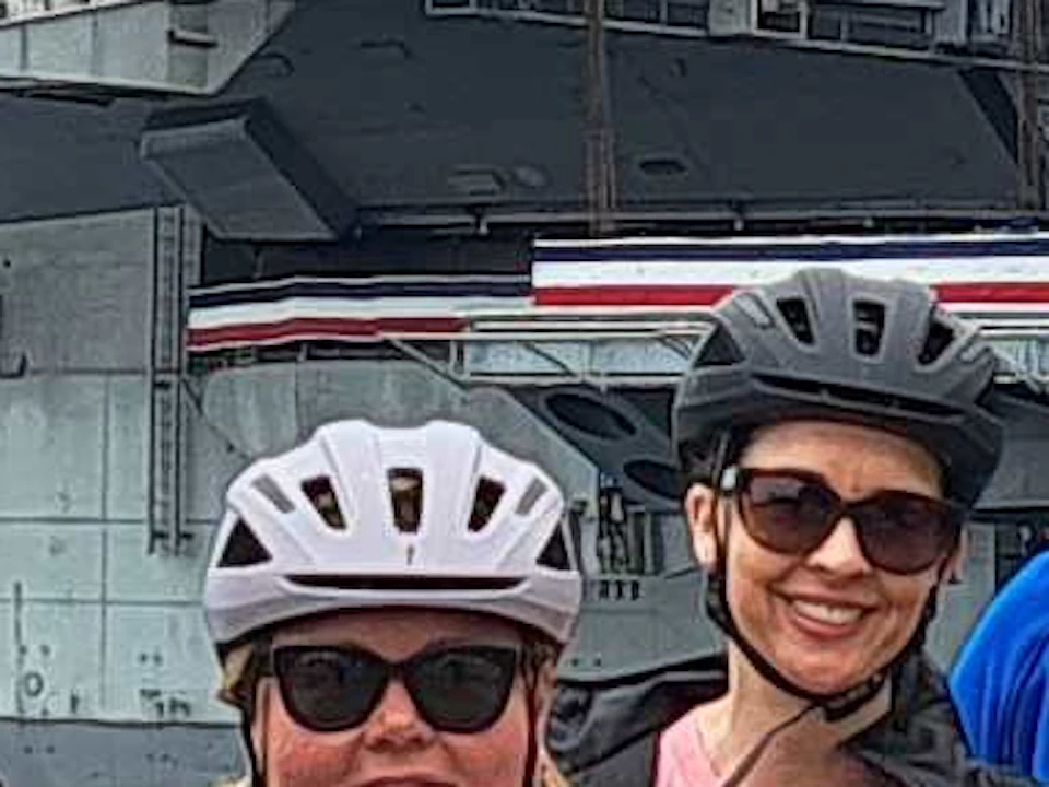 A group of people wearing helmets pose on a boardwalk with a large aircraft carrier docked in the background.