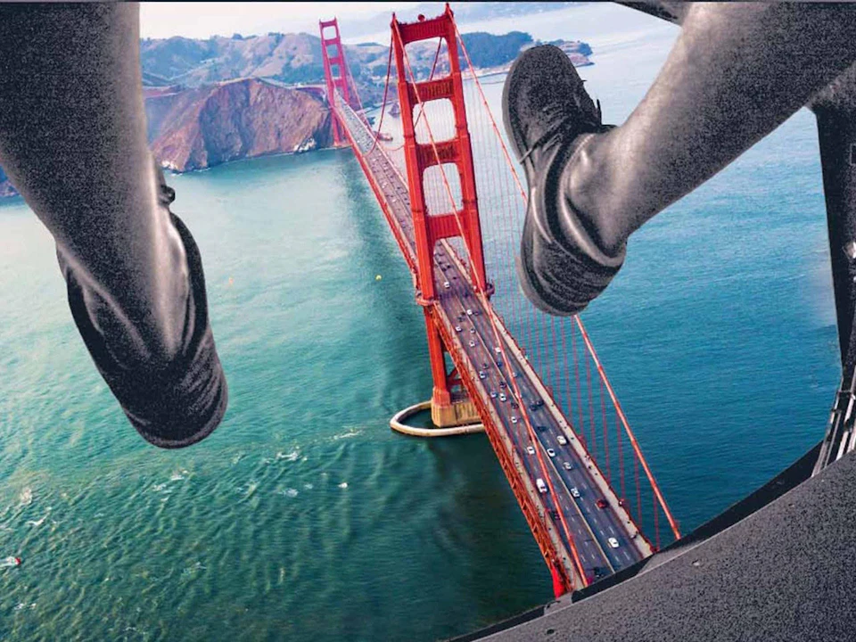 View of the Golden Gate Bridge from above, with a person's legs and shoes hanging out of a helicopter.