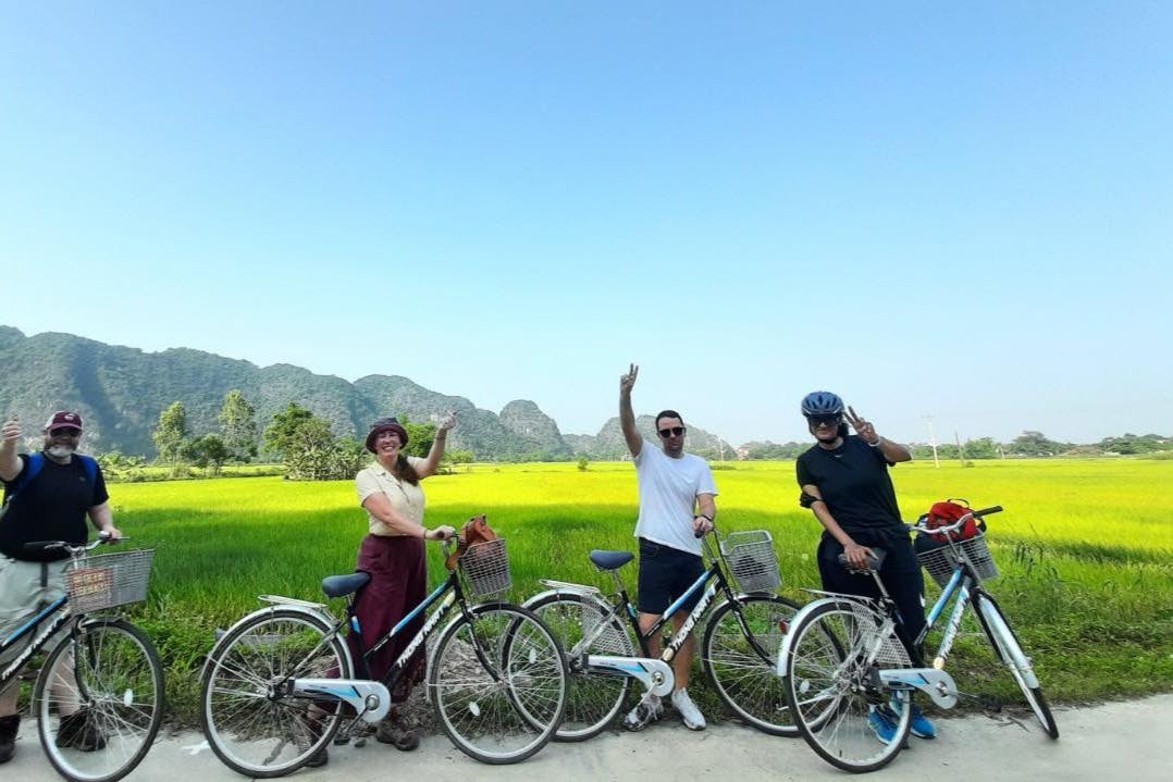 맑은 하늘 아래 푸른 들판과 먼 산을 배경으로 세 사람이 자전거를 타고 시골길에 서서 손을 흔들고 있습니다.