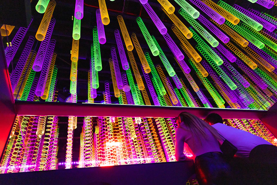 Two people leaning on a railing in a colorful space with hanging illuminated spiral decorations.