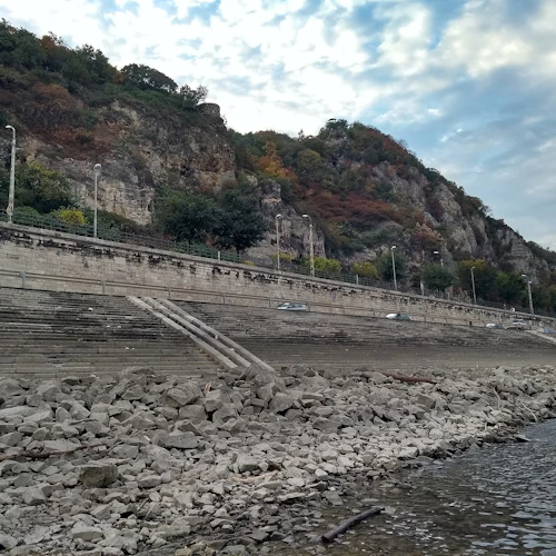 Stenig strandlinje med stentrappor som leder upp till en väg, flankerad av gatlyktor och träd, med en stenig kulle och molnig himmel i bakgrunden.