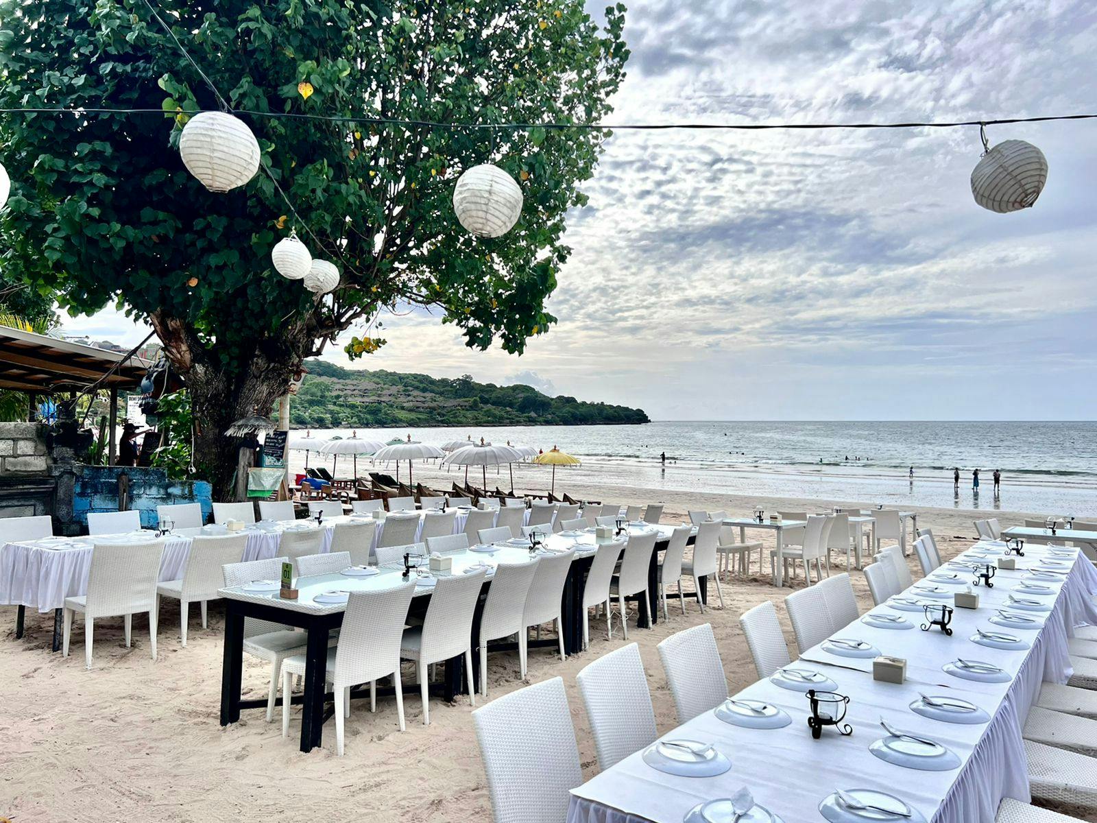 Jadalnia na świeżym powietrzu z białymi stolikami i krzesłami na piaszczystej plaży pod lampami sznurkowymi, w pobliżu oceanu w pochmurny dzień.