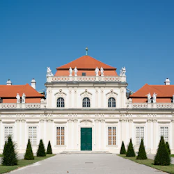 En historisk byggnad med röda tak, vita statyer och utsmyckade fönster, omgiven av välskötta koniska buskar och bänkar.