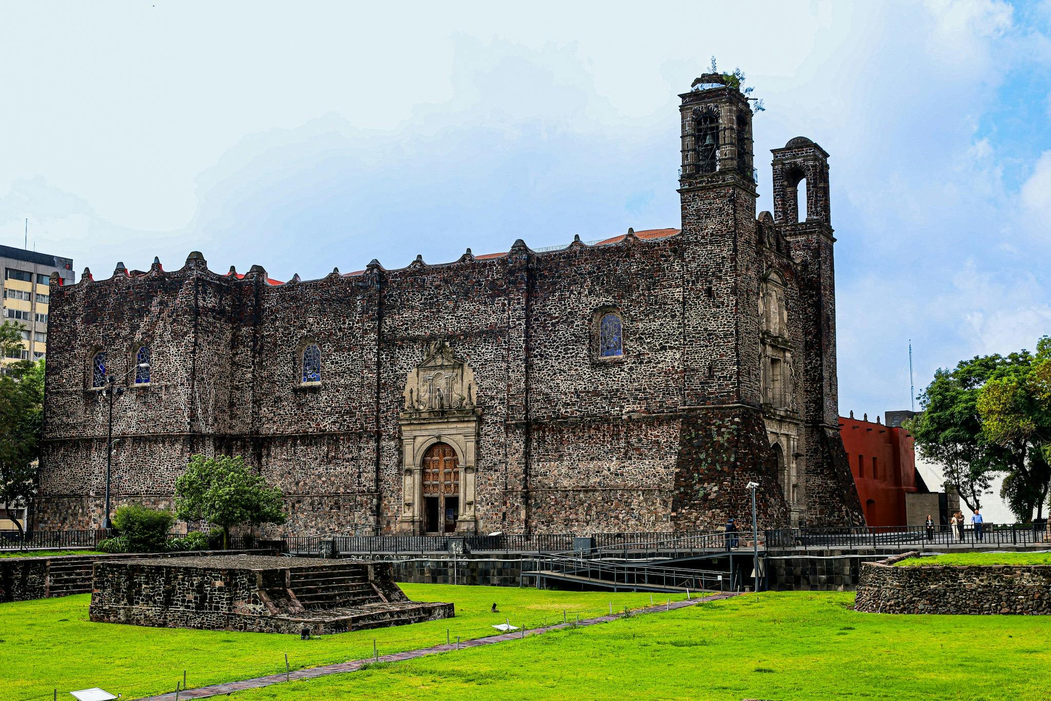 A historic stone church with tall walls and bell towers, surrounded by green grass under a cloudy sky.