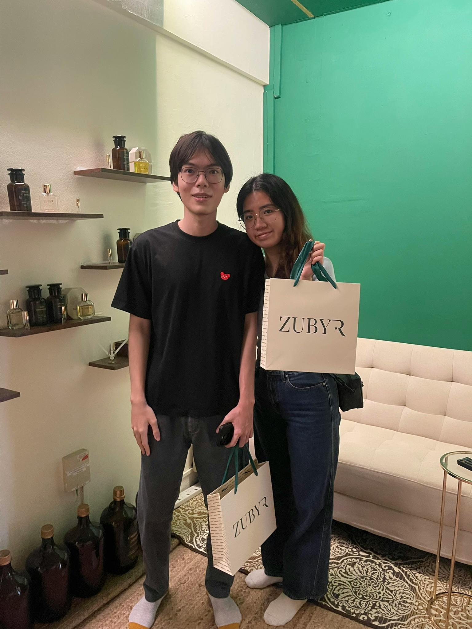Two people standing in a room with shelves containing bottles, one holding a shopping bag labeled 'ZUBYR.'