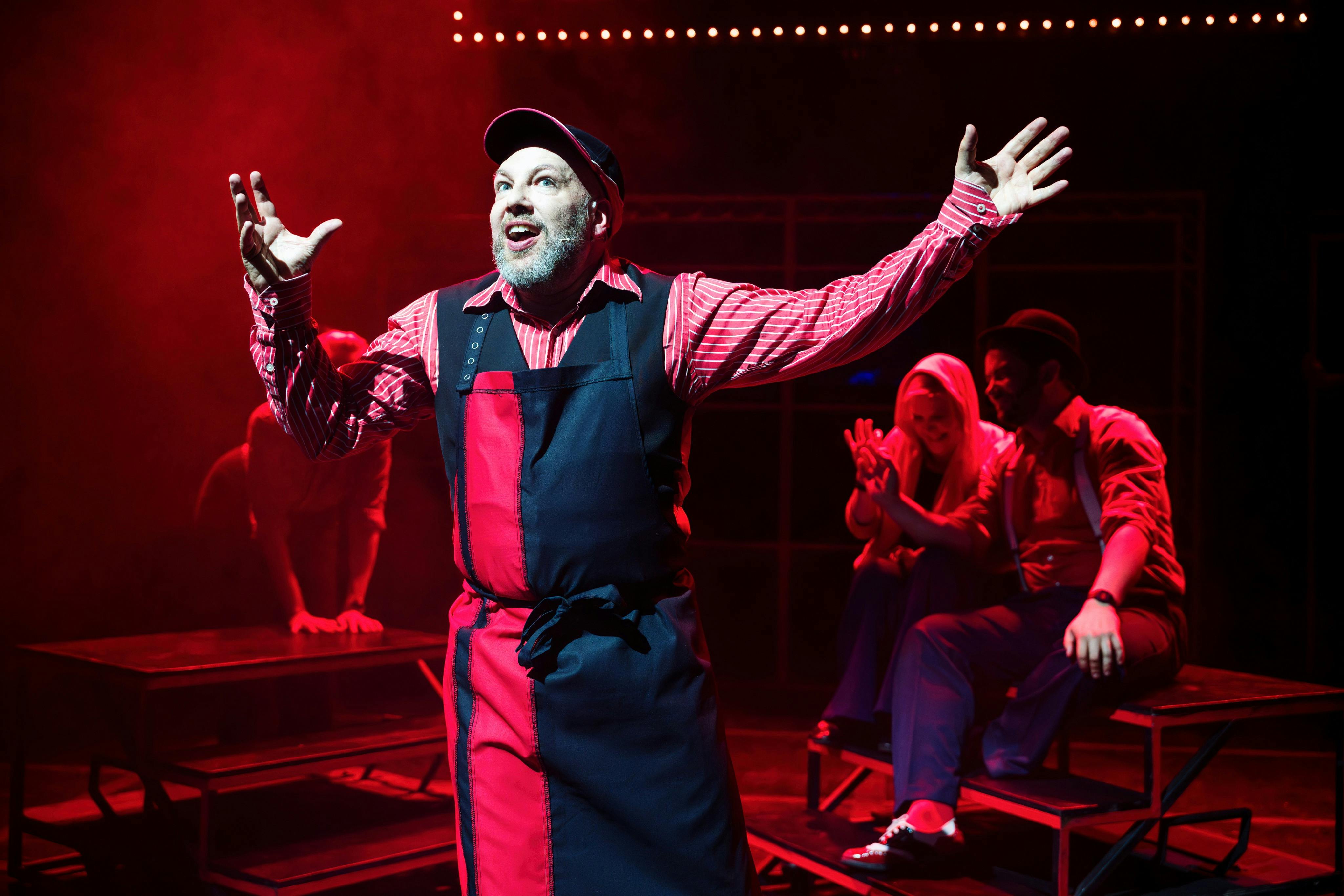 Vousatý muž v pruhované košili a zástěře vystupuje na jevišti pod červeným osvětlením. Za ním jsou vidět další účinkující.