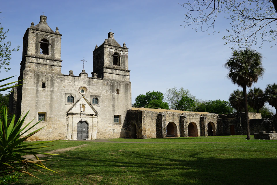 Historic stone church with twin bell towers and a row of arches, surrounded by green grass and trees under a clear sky.