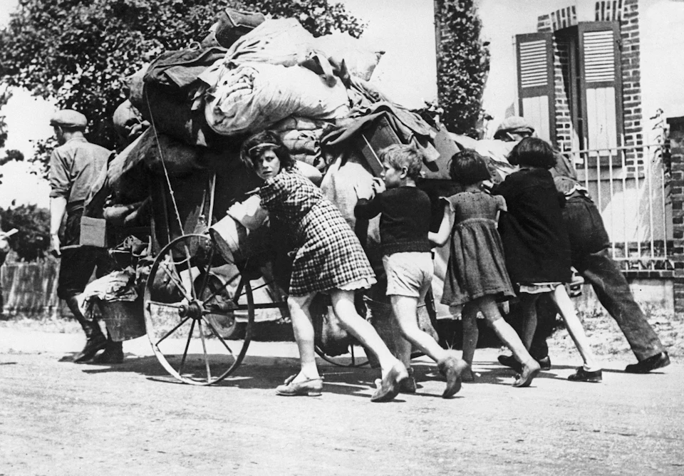 Children push and pull a cart overloaded with belongings down a street.