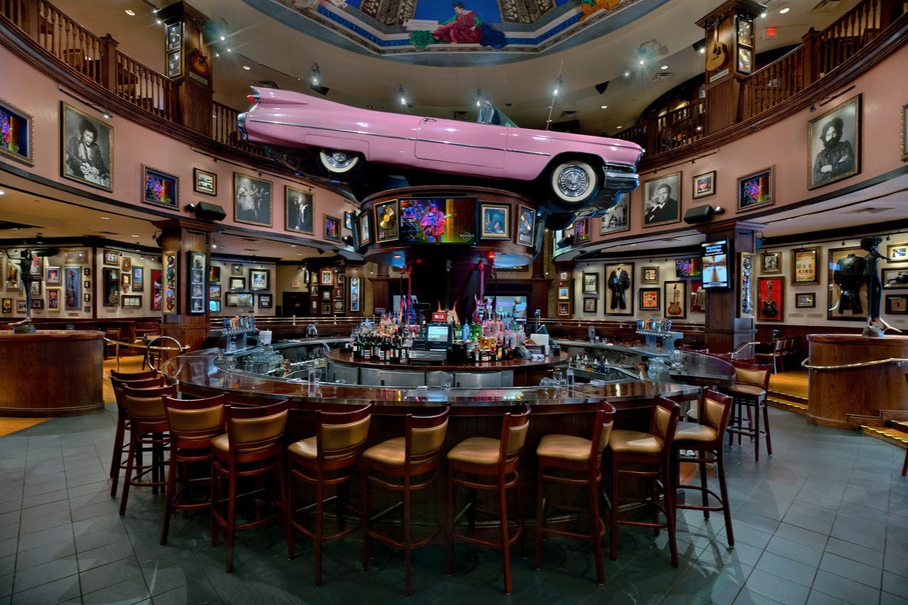 En bar med en lyserød bil over hovedet, omgivet af indrammede billeder og malerier, med tomme stole ved disken.