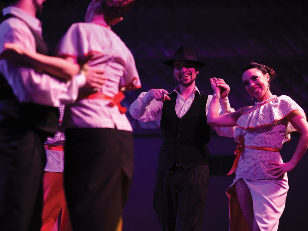 Dancers in formal attire performing on stage, illuminated in purple light. They hold hands and smile.