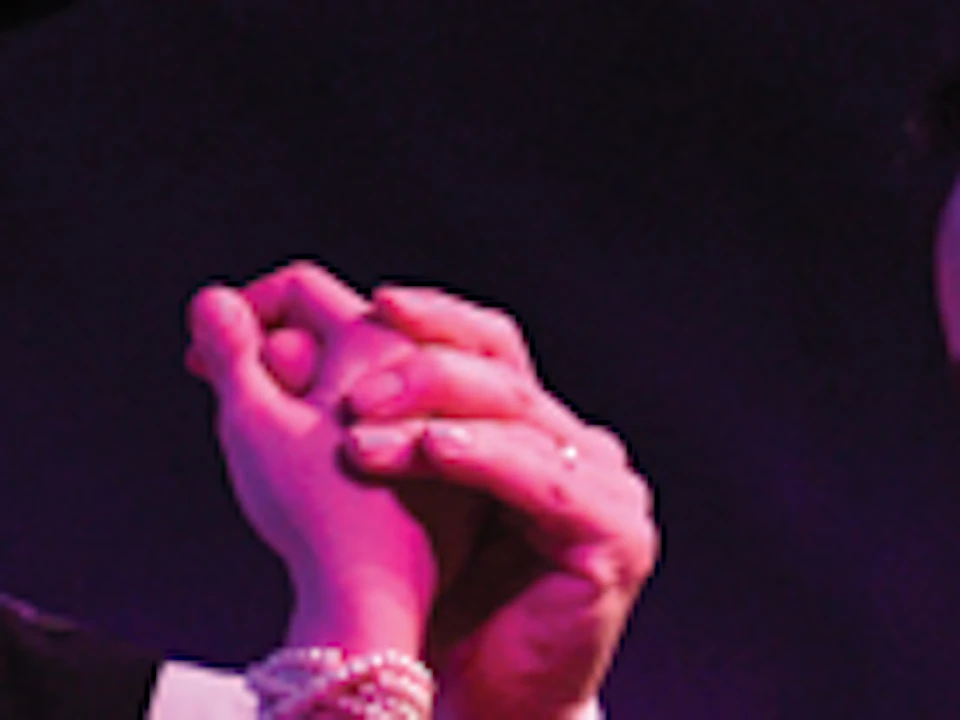 Dancers in formal attire performing on stage, illuminated in purple light. They hold hands and smile.