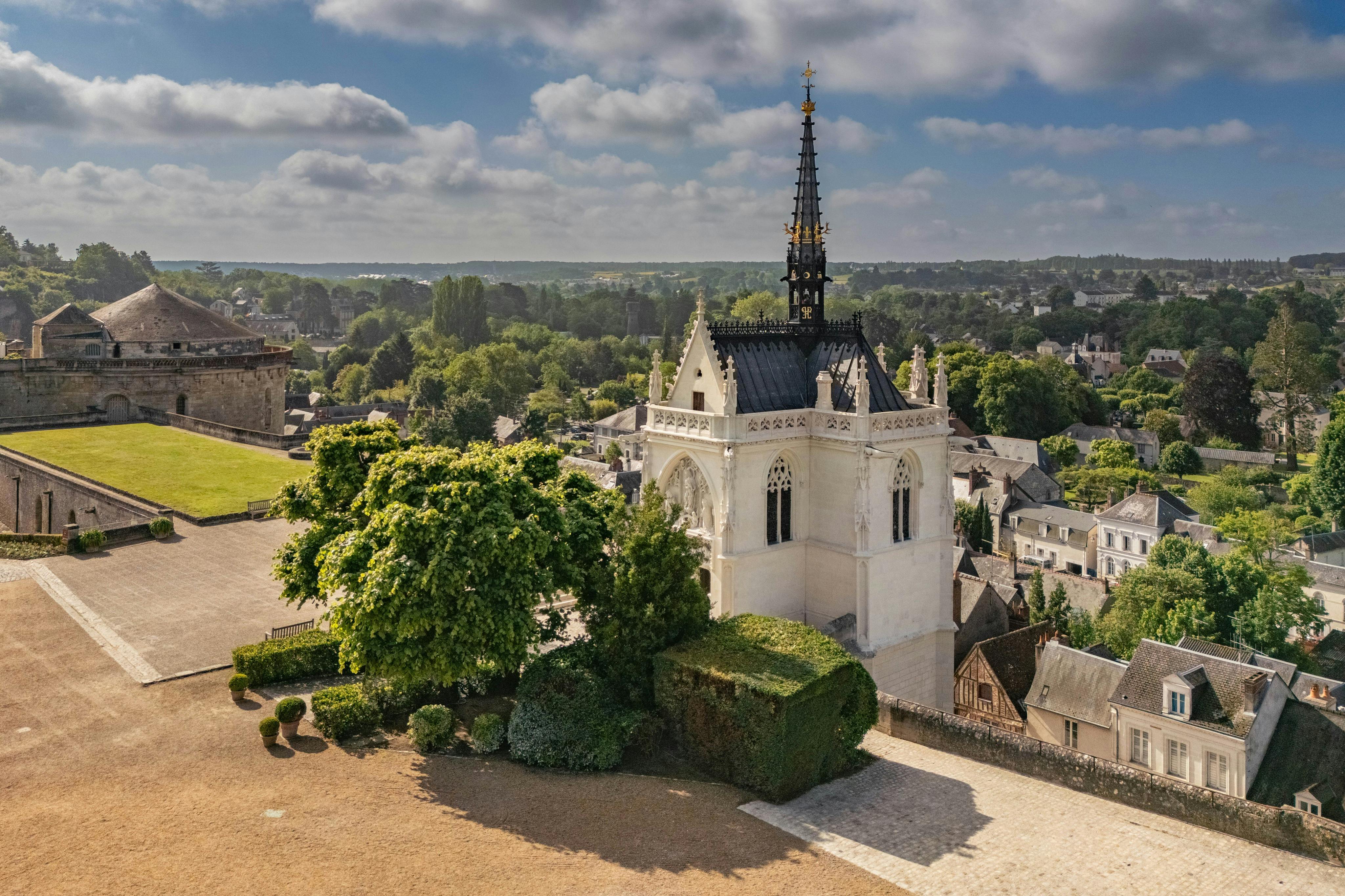 Una capilla histórica con una alta aguja rodeada de vegetación y con vistas a una ciudad bajo un cielo parcialmente nublado.